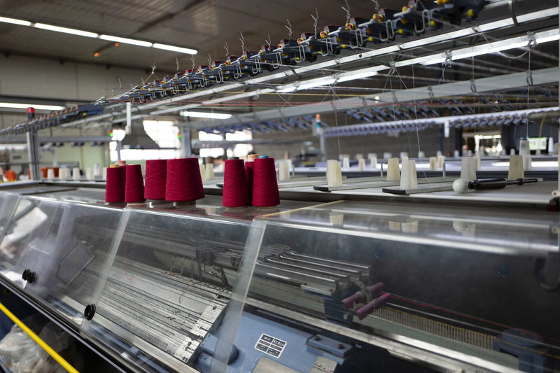 Industrial flat knitting machines lined up inside BENTA knitwear OEM factory workshop, red yarn cones placed on production equipment.